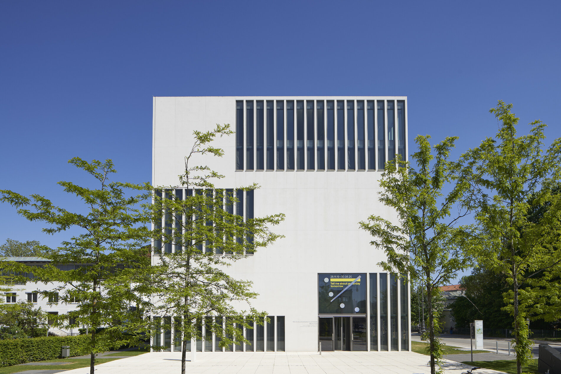 Будівля у формі білого кубу з вузькими видовженими вікнами, перед нею простора площа і дерева 