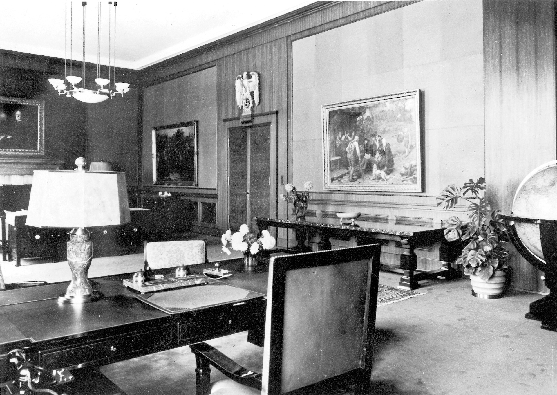 A room with a large desk, seating around a fireplace, and several paintings on the wall.