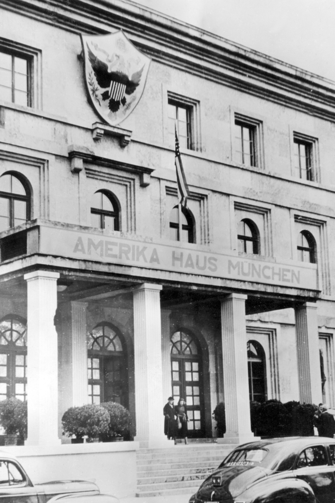 Blick von der Straße auf die Frontfassade eines dreistöckigen Gebäudes Über dem Säuleneingang steht „Amerika Haus München“, darüber hängt die Flagge der USA.