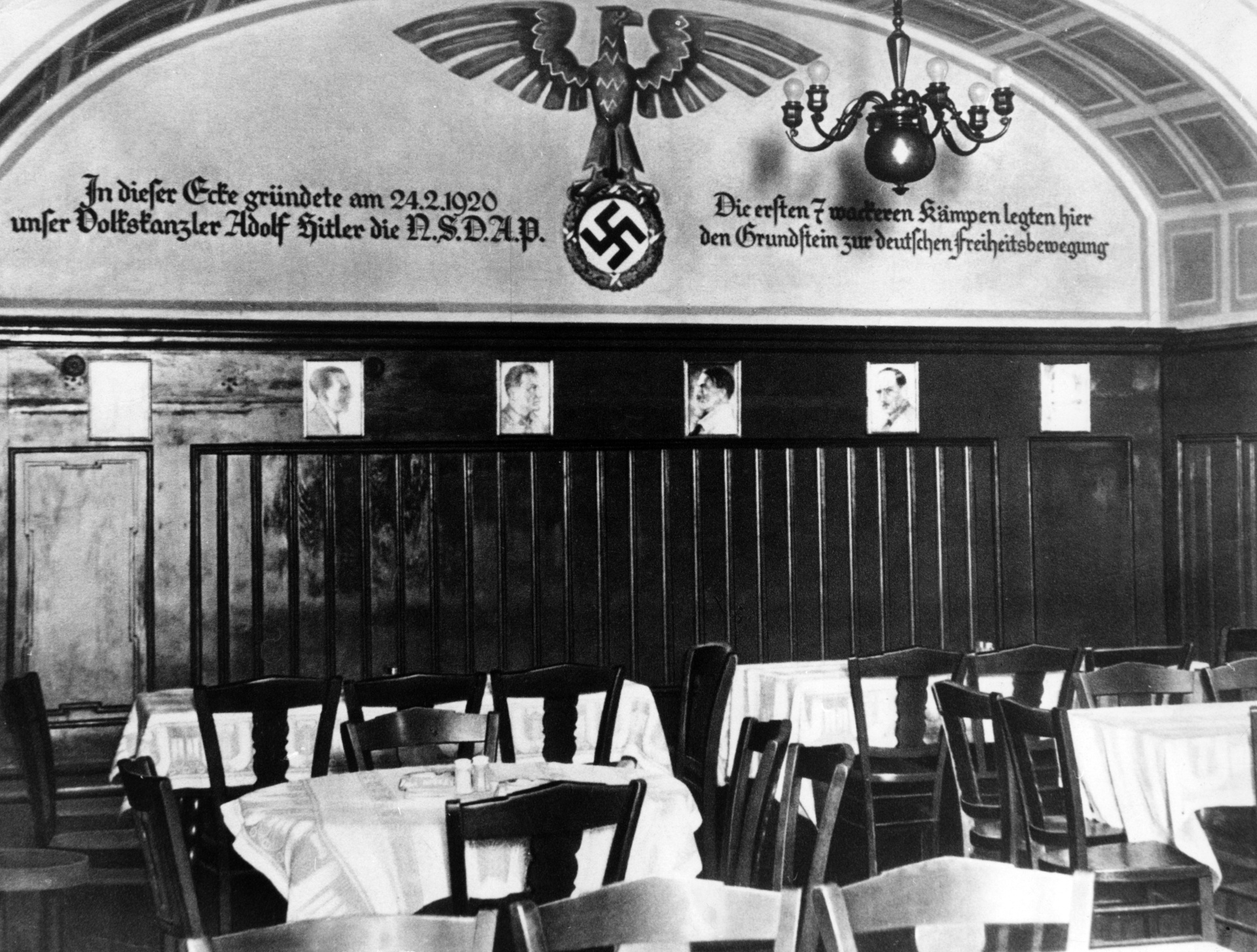 Hinter Tischen und Stühlen hängt an einer Wand ein Hakenkreuz. Daneben steht ein Text zur Gründung der NSDAP. 