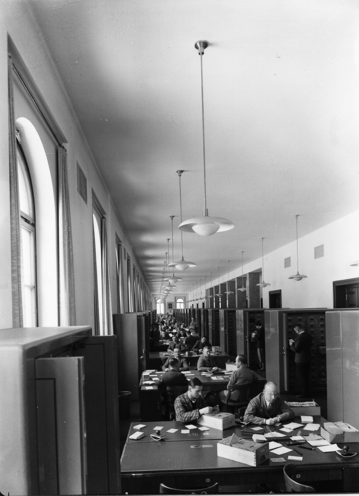 A long corridor with tables arranged one behind the other at which a number of men are sorting files. Cabinets stand to their left and right.