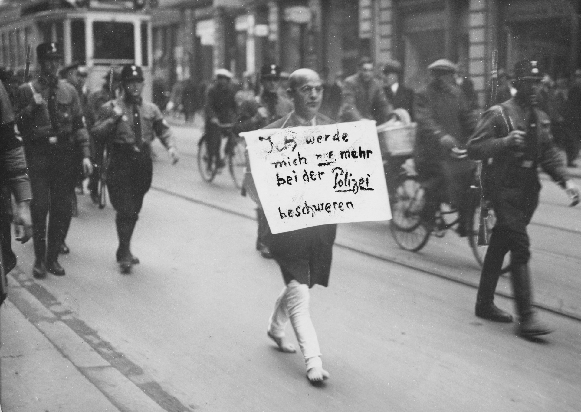 A man walking barefoot along the street with cut-off pants and a sign around his neck saying: “I’ll never complain to the police again.” Behind and beside him are men in uniform and passers-by.