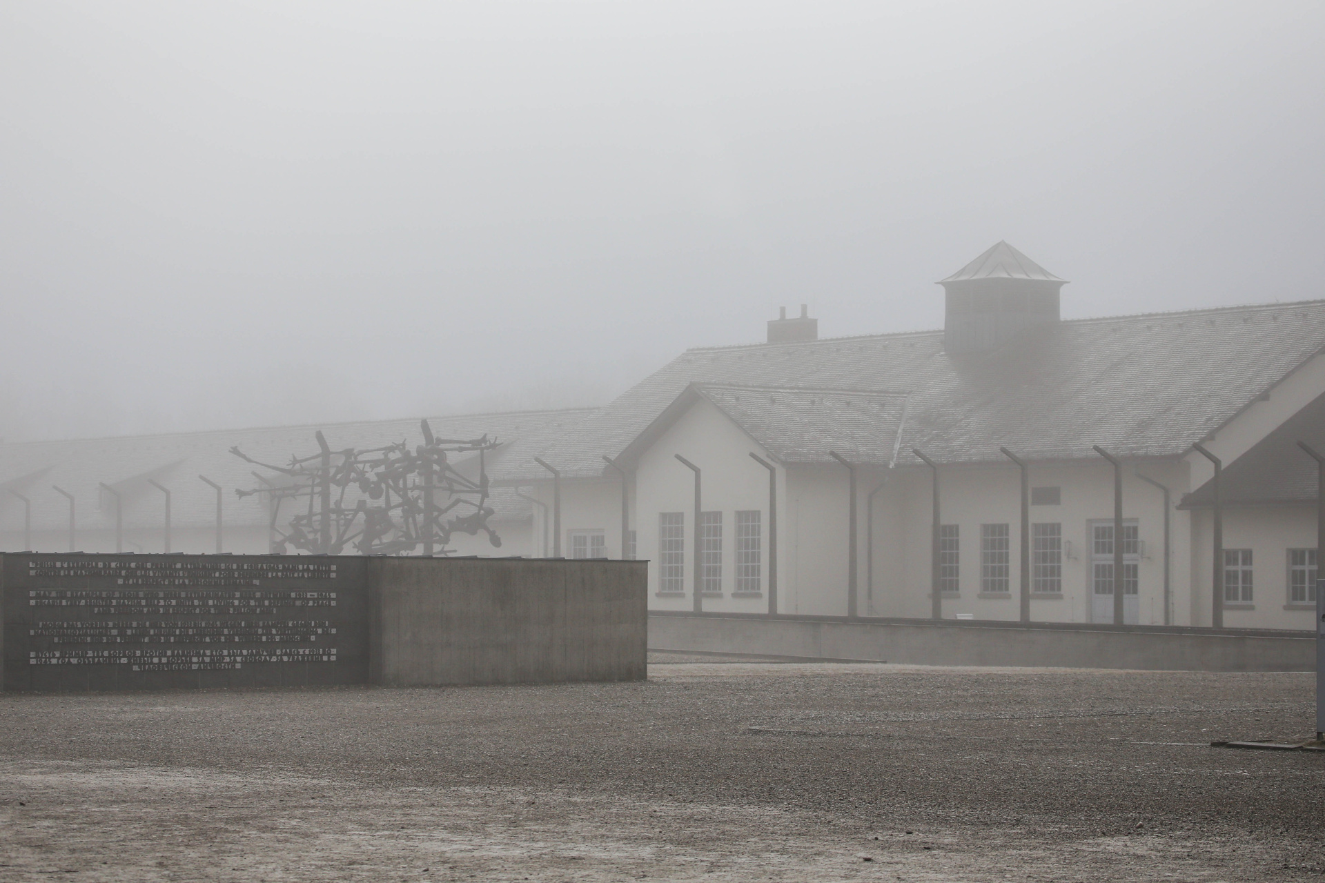 A building stands behind a fence in the fog. In front of it is a wall and a large open space.