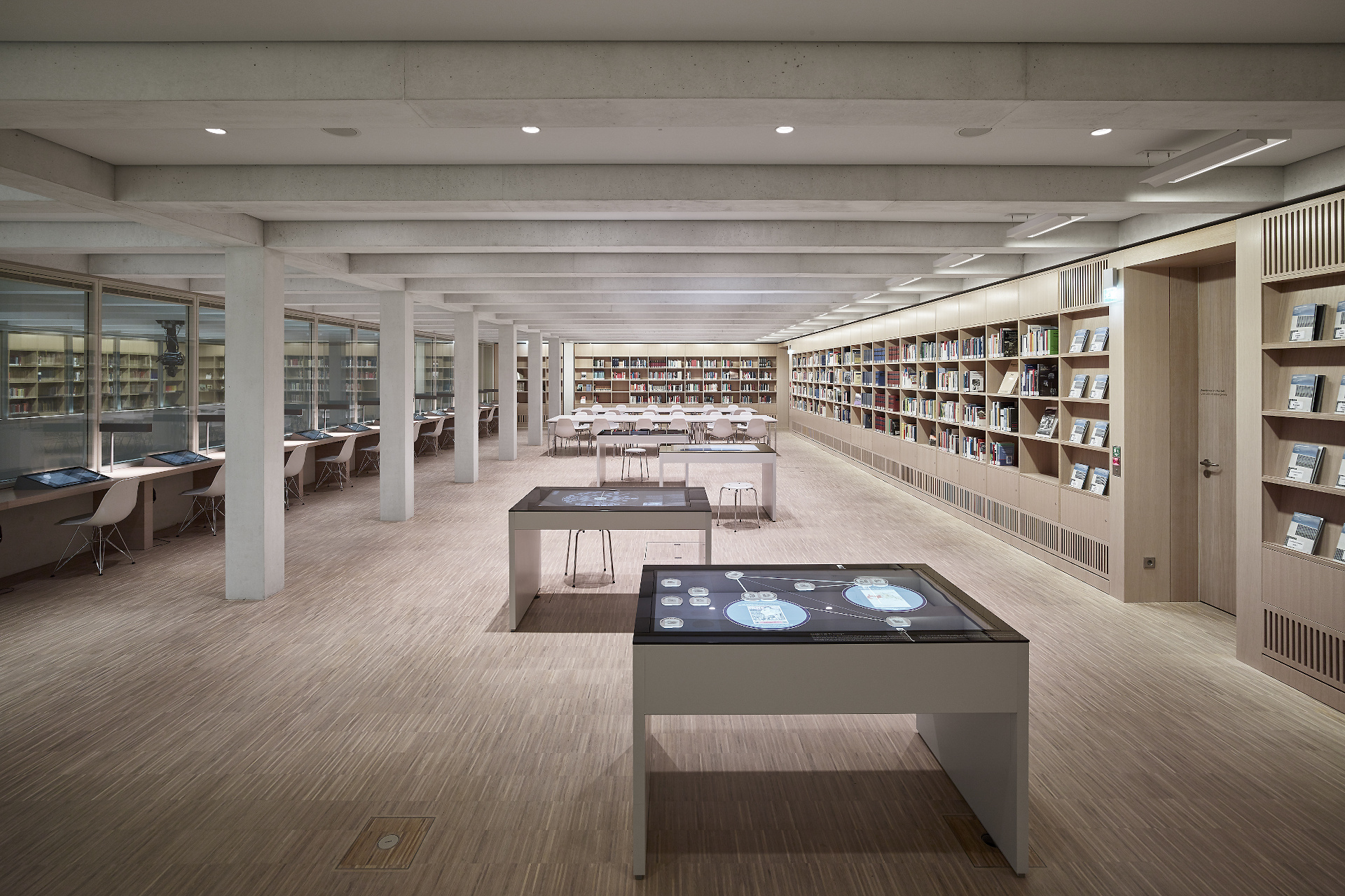 In the foreground are four media tables. On the right are touch screen terminals and on the left the bookshelves in the reference library. 