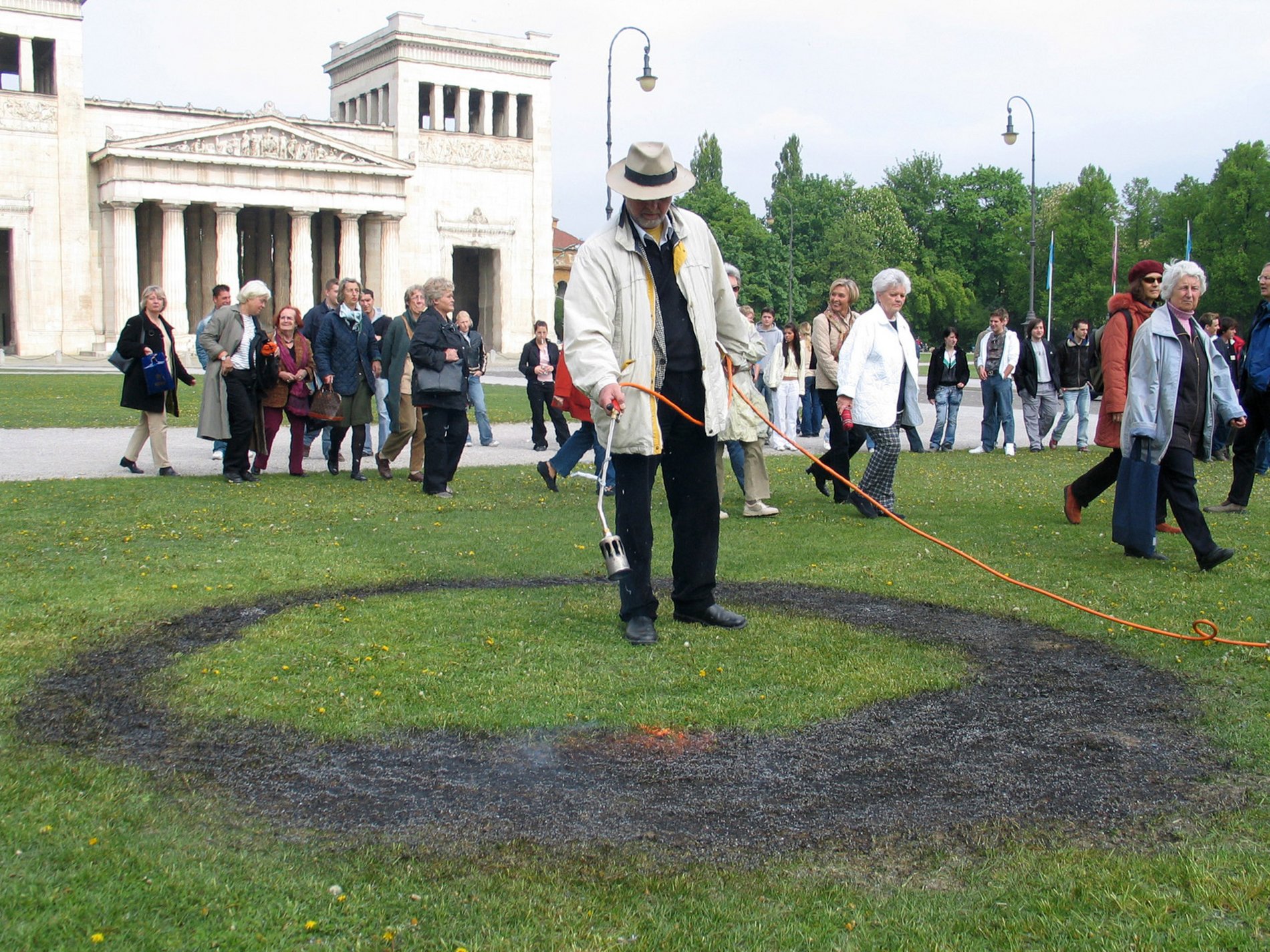Mit einem Bunsenbrenner brennt ein Mann umgeben von mehreren Zuschauer*innen einen kreisrunden Brandfleck in die Wiese vor den Staatlichen Antikensammlungen.