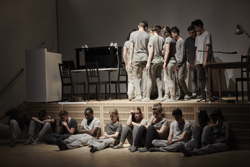 In grau gekleidete Schüler*innen stehen in einer Reihe hintereinander auf der Bühne im Auditorium des NS-Dokumentationszentrums. Einige sitzen vor der Bühne und blicken auf den Boden.