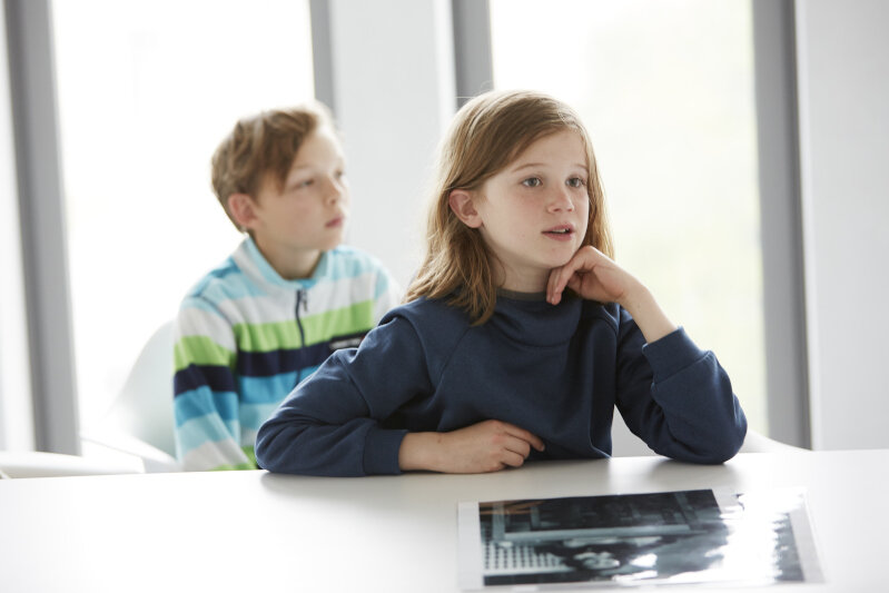A girl and a boy are sitting at a table in a meeting room at the Munich Documentation Center. A laminated photograph is lying on the table in front of them.
