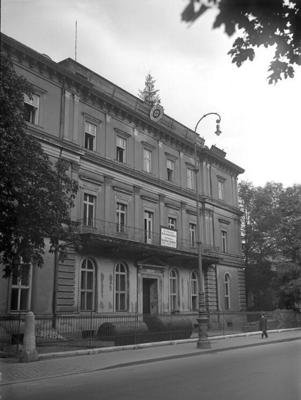 View from the street of the facade of a three-storey building with many windows. A sign with a swastika hangs from the roof.