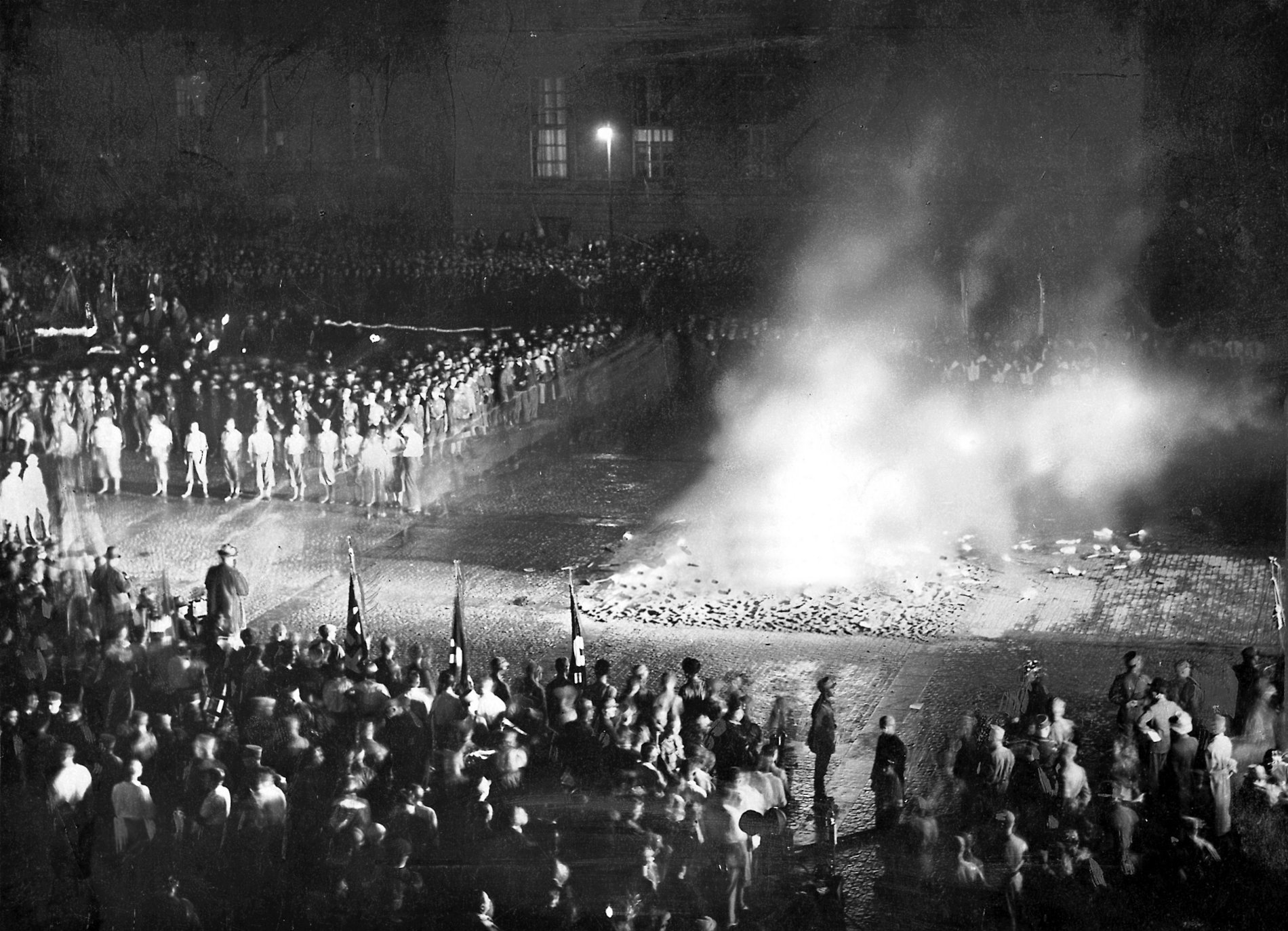 Tausende nationalsozialistische Studierende und SA-Männer stehen teils mit Fahnen um ein loderndes Feuer auf dem Berliner Opernplatz, in dem Bücher verbrennen. 
