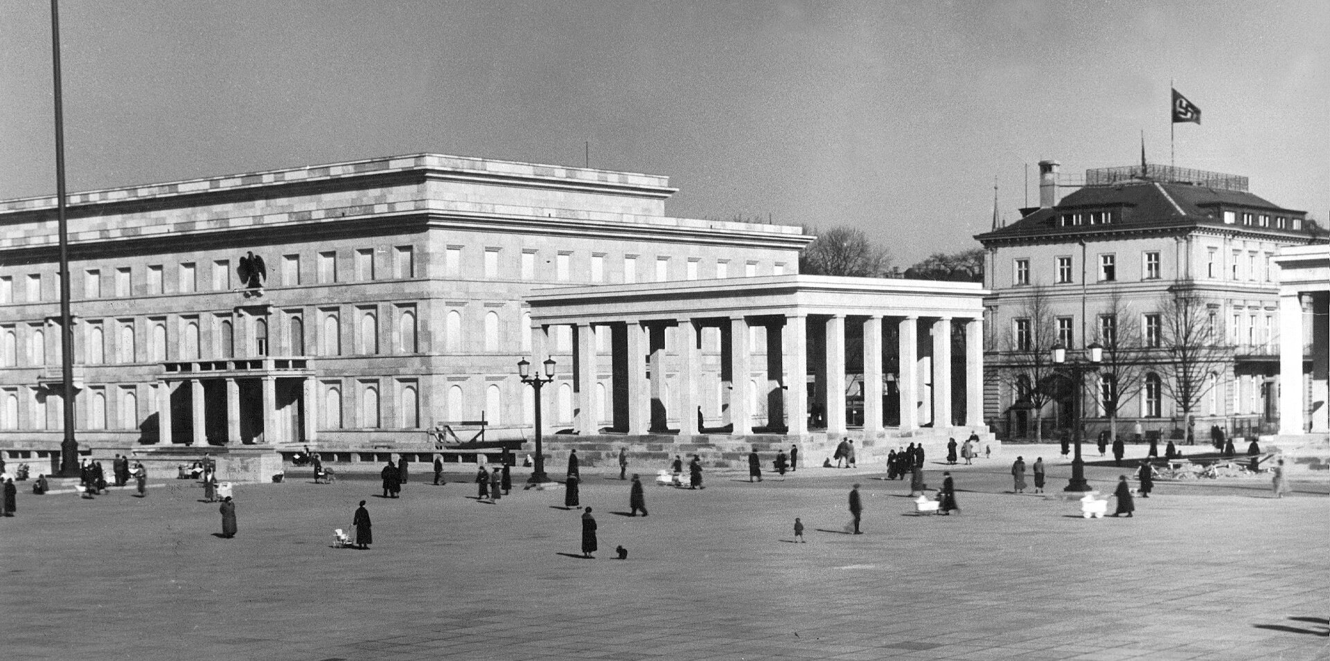 A square paved with granite slabs. On the right is a long building, in the middle a hall of columns, and on the right a building with a swastika flag. People are walking across the square. 