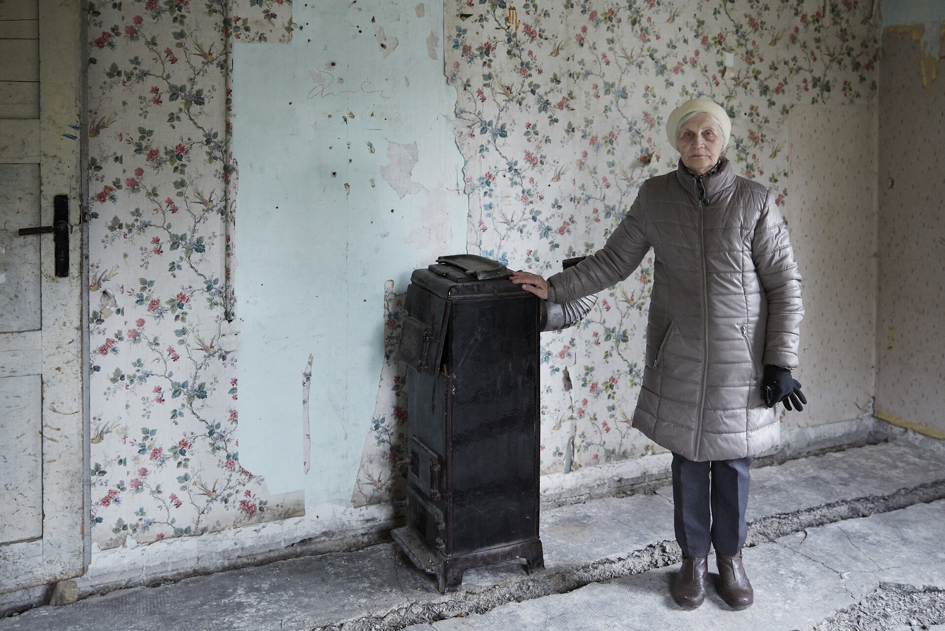 Eine ältere Frau steht in einem Raum mit Blumentapete. Mit einer Hand stützt sie sich an einem alten Ofen.