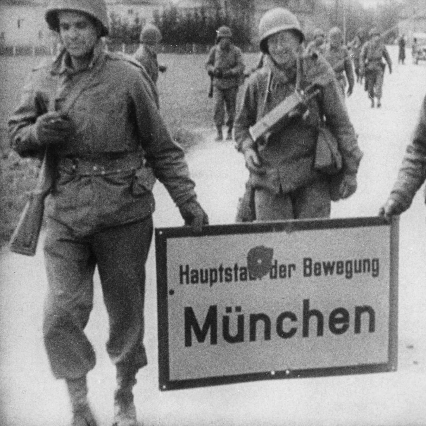 Three armed American soldiers carrying a place-name sign with a bullet hole in it. The sign reads “Munich, Capital of the Movement.”