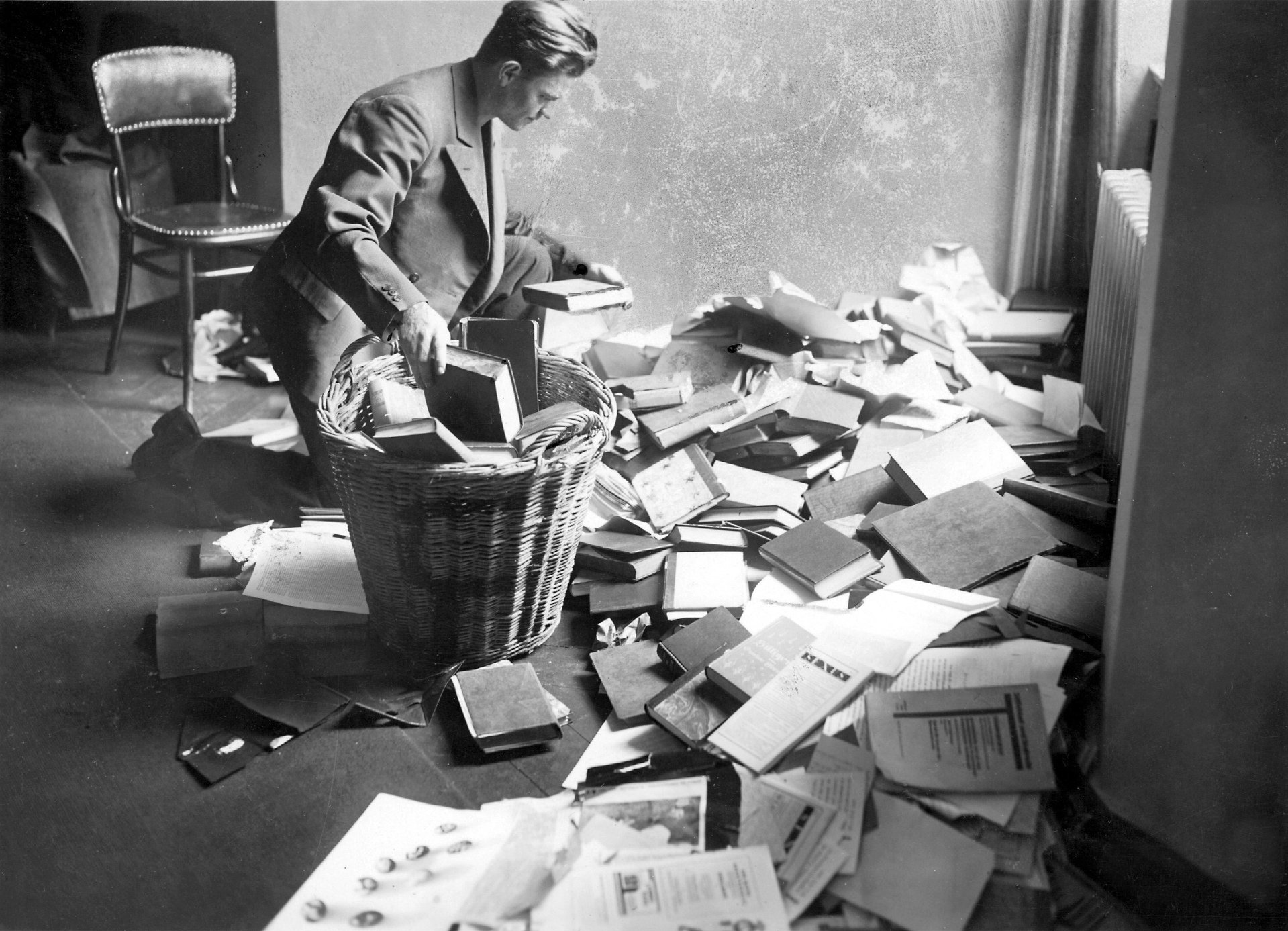 A student standing in a room in front of a pile of books and writings that have been thrown on the floor. He is picking them up and throwing them into a large trash basket.