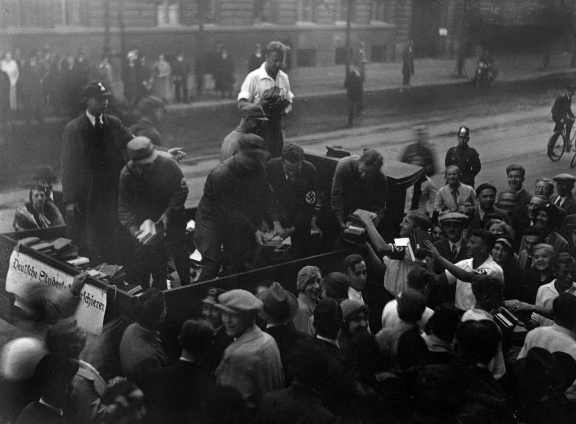  Pro-Nazi students are standing on the loading platform of a small open truck at the edge of the road and loading books brought by a crowd of people into it.