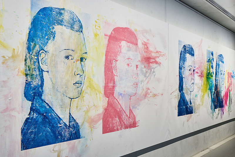 Gemälde mit fünf bunten Frauenköpfen an einer weißen Wand 