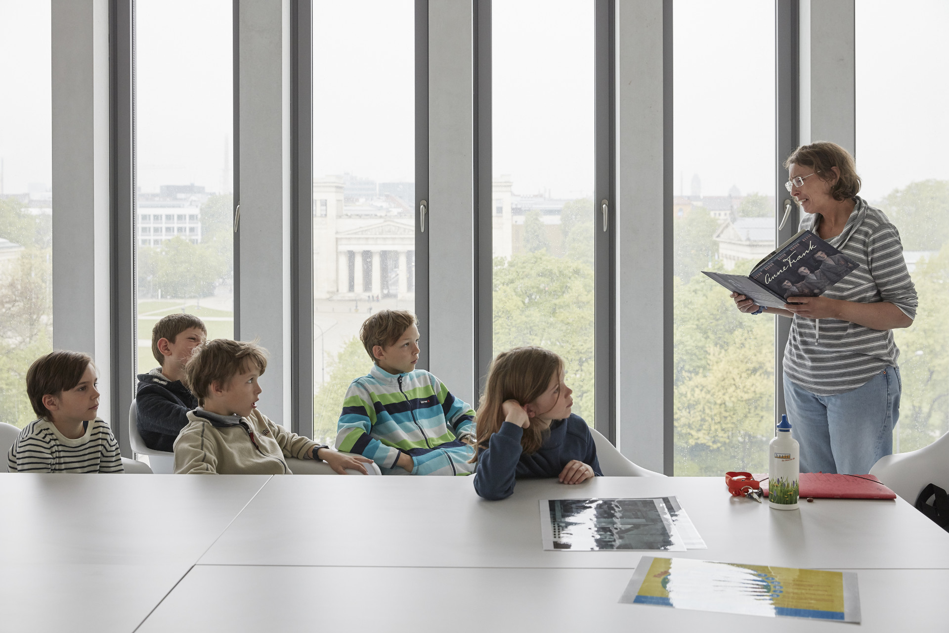 Five children are sitting at a table in a meeting room at the Munich Documentation Center. A member of staff is standing in front of them reading aloud from a book.