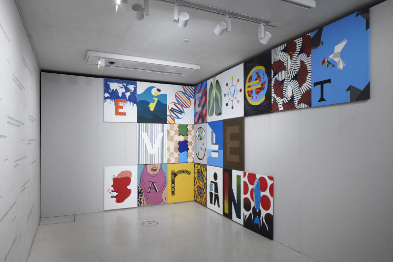 Several colorfully painted and decorated wooden panels hang side by side on a wall. Each panel shows one letter of the slogan “One world, many colors.”