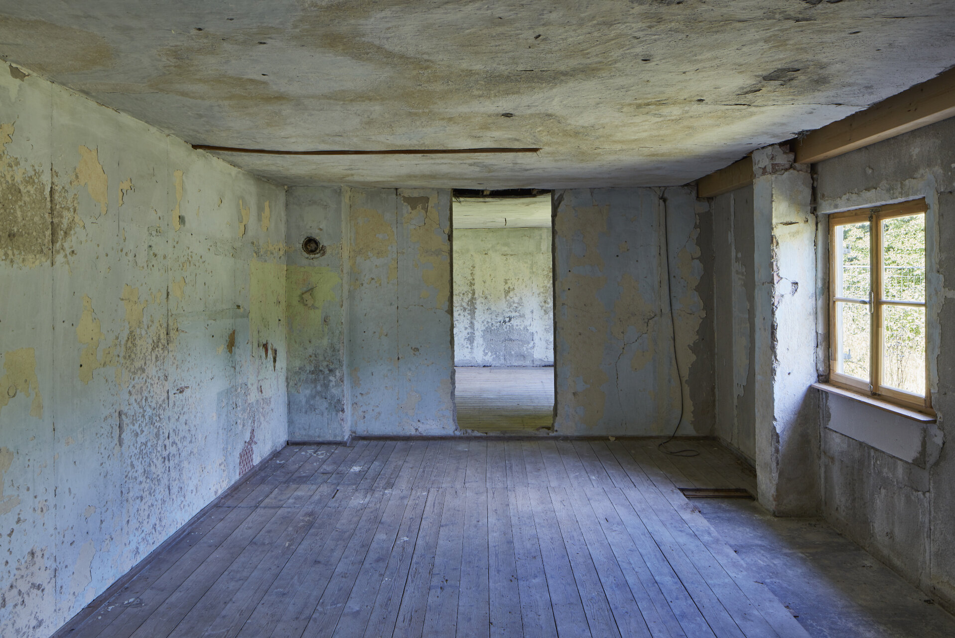 Two rooms with bare concrete walls inside the empty barrack. The room has windows but no doors.  