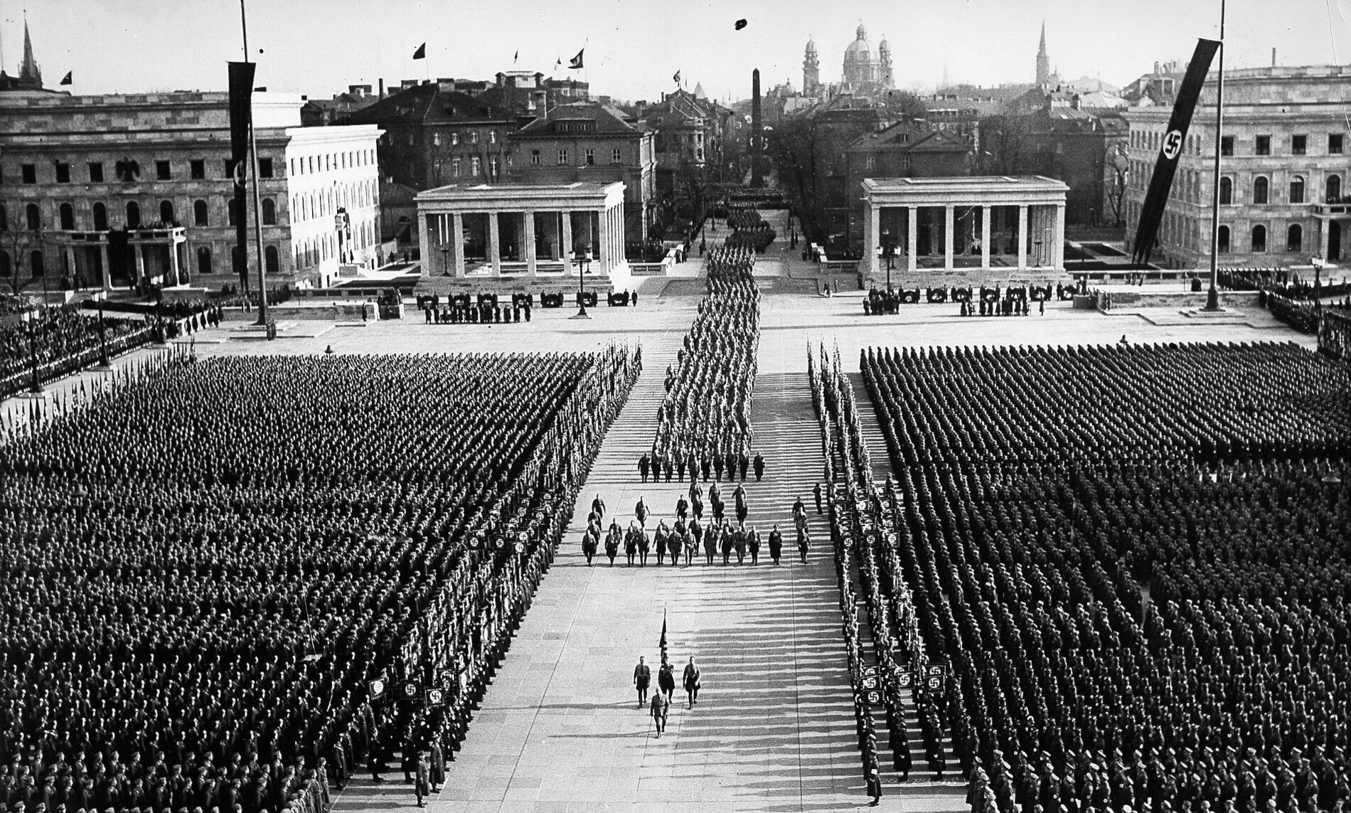 View from the Propylea onto Königsplatz adorned with swastikas. A funeral procession led by flag-bearers is making its way down the center of the square flanked on either side by hundreds of people in uniforms.