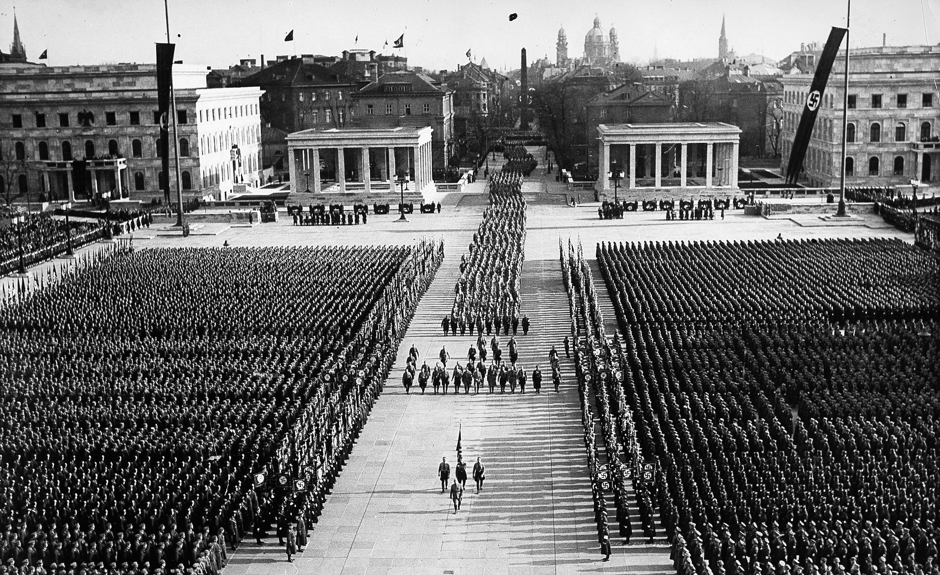 [Translate to English:] Gamalı haç bayraklarıyla süslenmiş Königsplatz'ın ortasından, yüzlerce üniformalı adamın sıralandığı, bayrak taşıyıcılarının önderlik ettiği bir cenaze alayı  geçiyor.