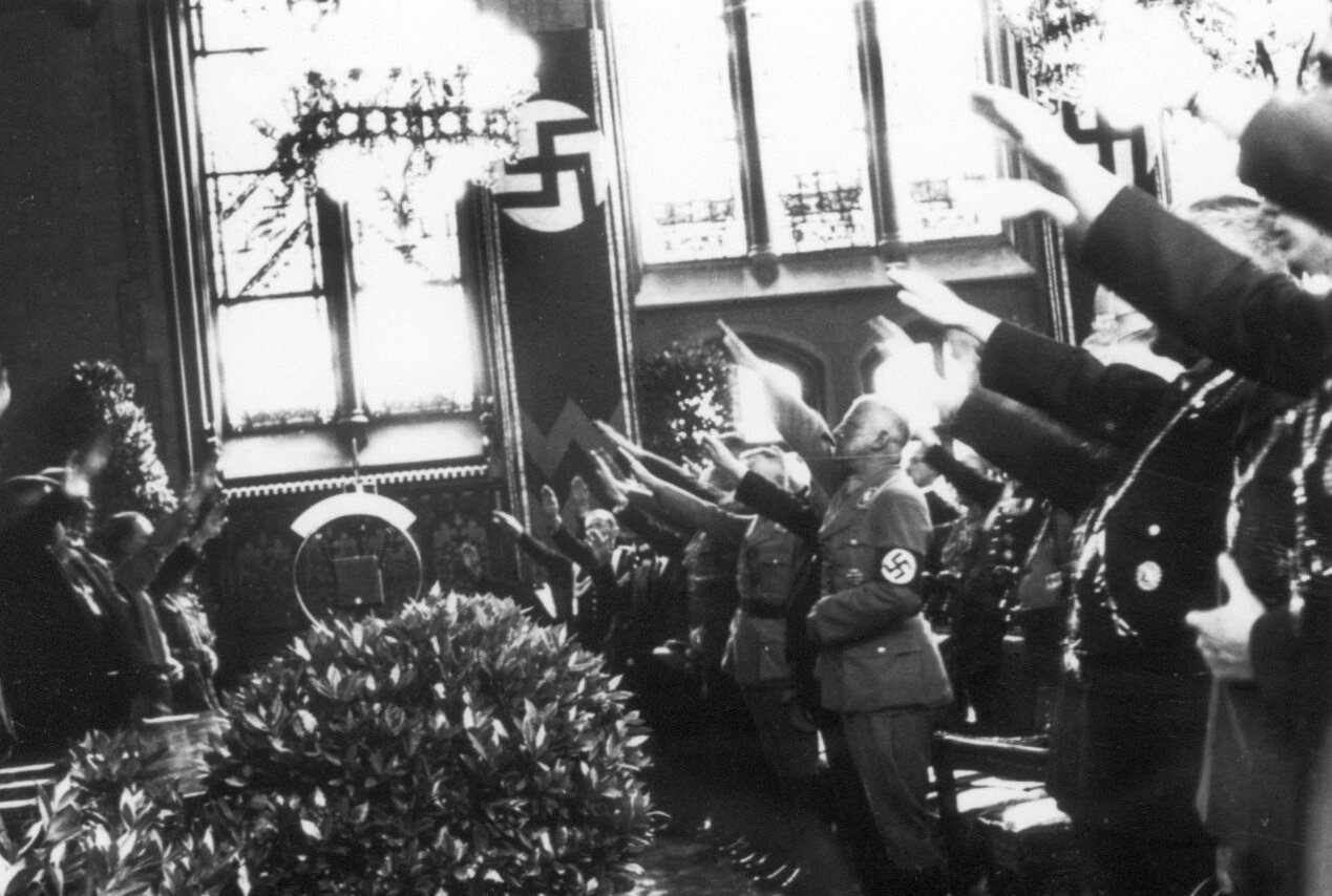Zahlreiche uniformierte Männer stehen im Münchner Rathaus nebeneinander und heben die Hand zum Hitlergruß.