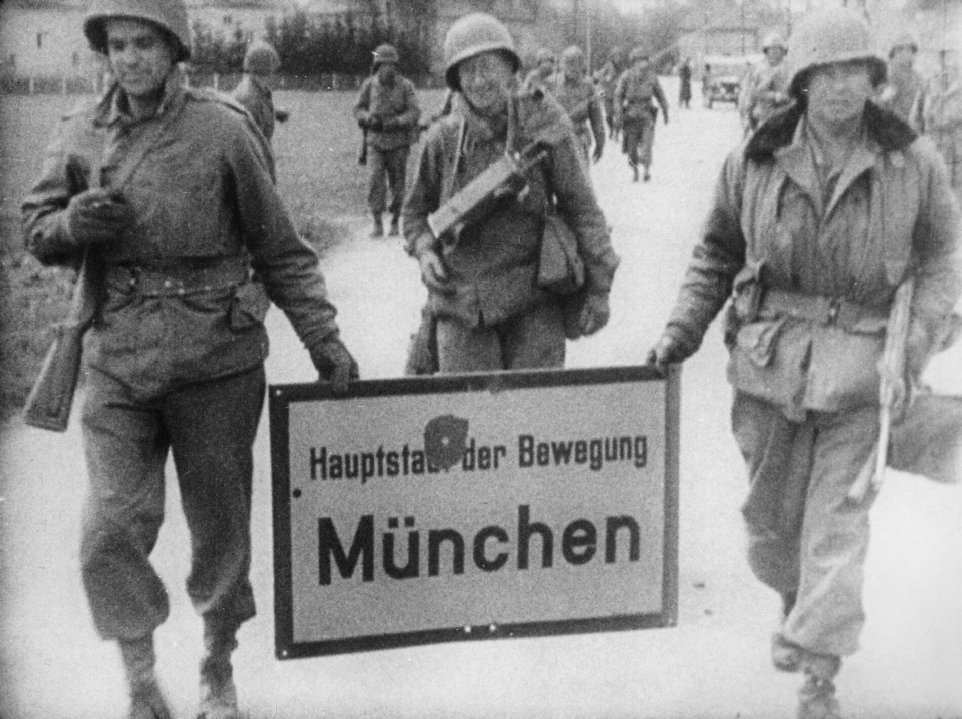 Drei bewaffnete amerikanische Soldaten tragen ein Ortsschild mit der Aufschrift „Hauptstadt der Bewegung München“ und einem Einschussloch.